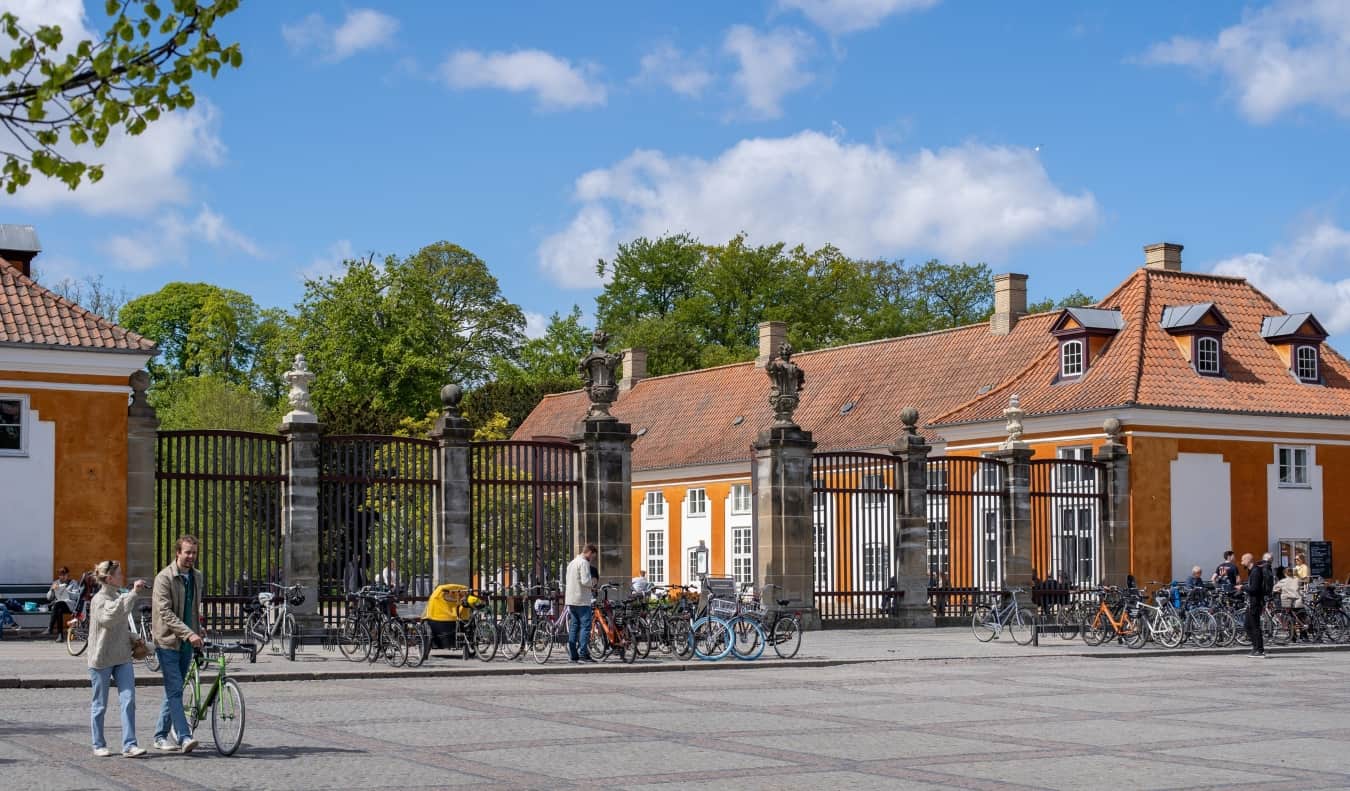 People walking by a large gated entrance to a tree-filled park, flanked by historic orange buildings in Copenhagen, Denmark