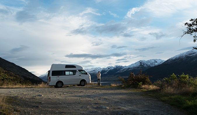 man stood next to a van in New Zealand in the background snowcapped mountains